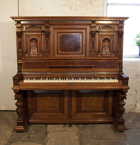 A German upright piano with a Neoclassical style walnut case and cup and cover legs. Cabinet features ornately carved pilasters in high reief and copper sconces in a sea monster design. Piano has an eighty-five note keyboard and two pedals 